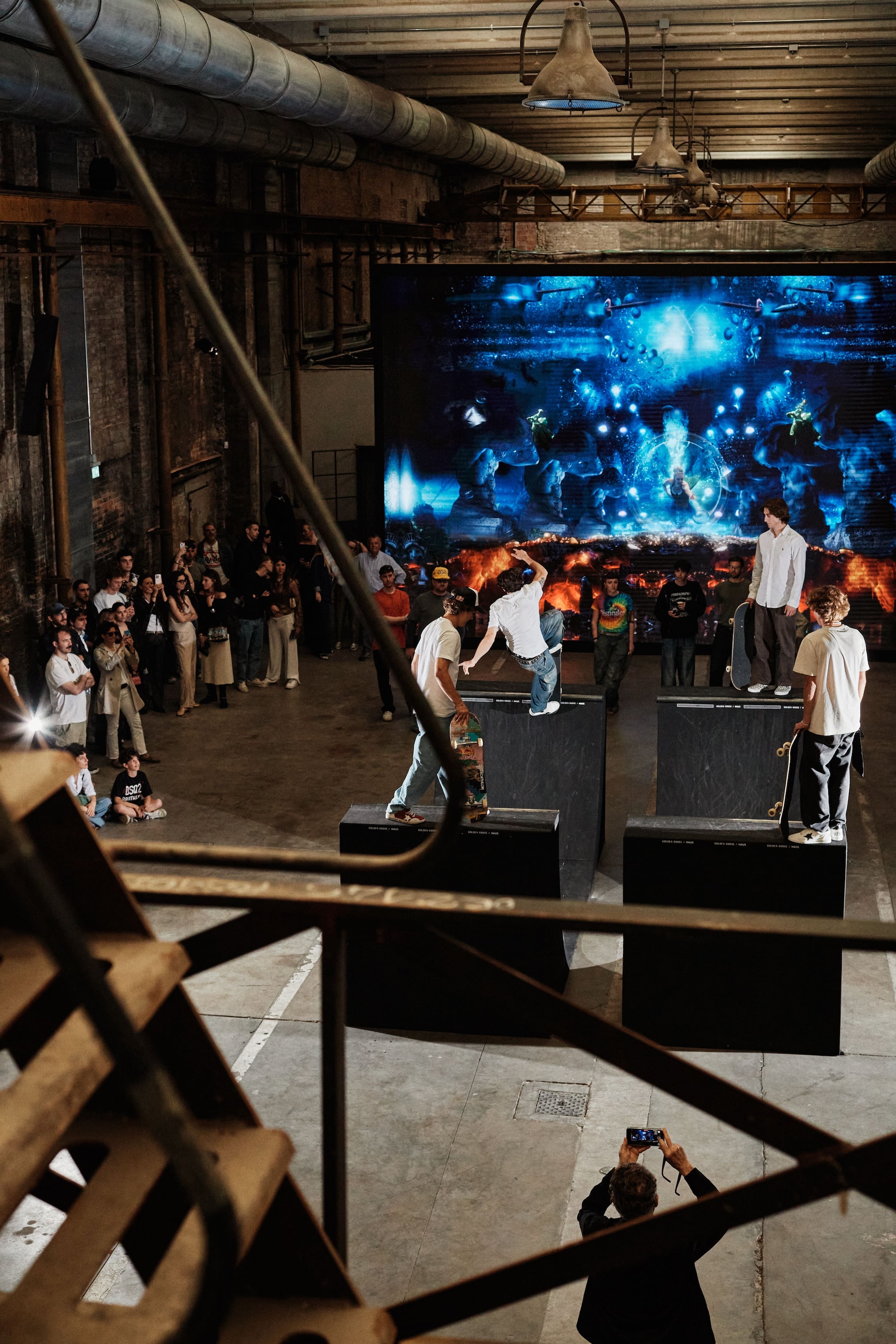 Skateboarders performing tricks on ramps in front of large digital screen at Haus event